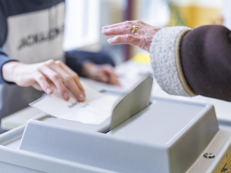 Stimmabgabe in einem Wahllokal bei der Bundestagswahl: Eine Hand wirft den Wahlschein in die Urne, die von einer anderen Hand freigegeben wird.