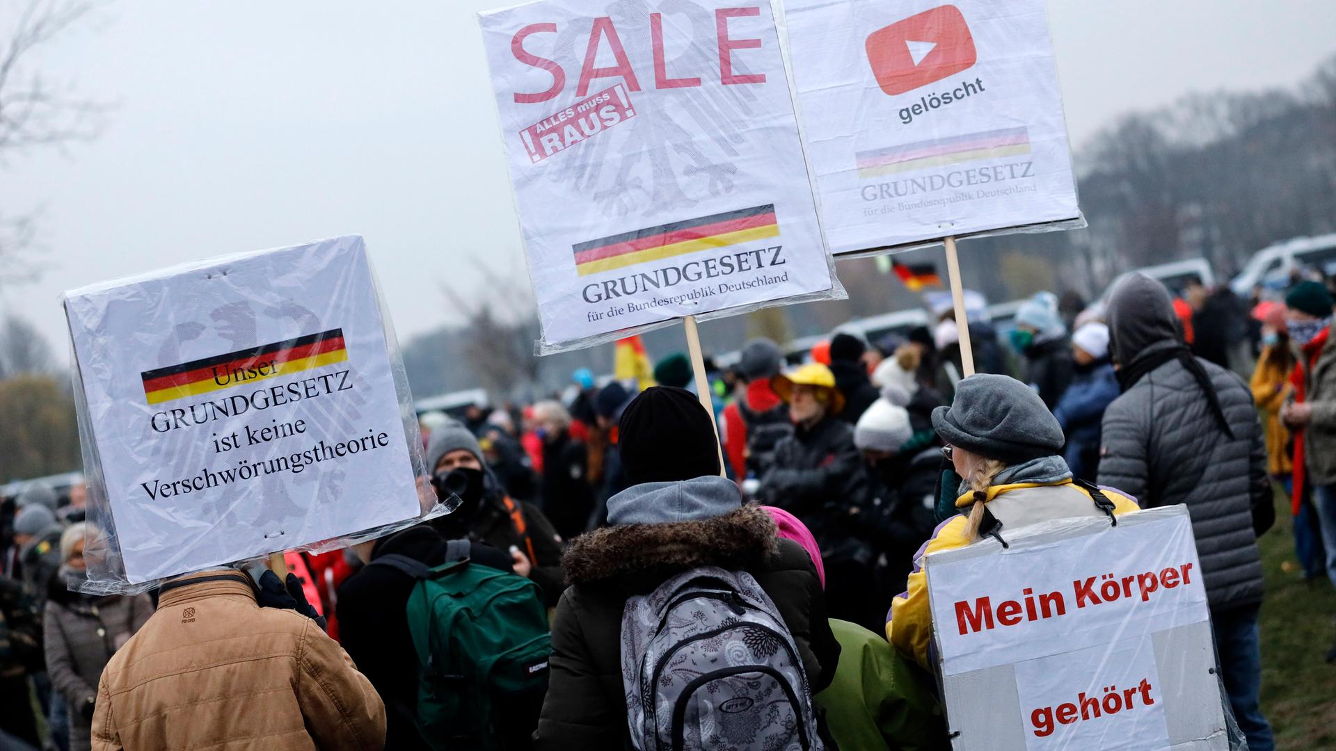 Mehrere Personen stehen bei einer Querdenken-Demonstration im Freien. Sie halten Plakate mit Aufschriften wie „Mein Körper gehört mir“ und „Grundgesetz ist keine Verschwörungstheorie“. Im Hintergrund ist eine Deutschlandfahne zu sehen. Mehrere Personen stehen bei einer Querdenken-Demonstration im Freien. Sie halten Plakate mit Aufschriften wie „Mein Körper gehört mir“ und „Grundgesetz ist keine Verschwörungstheorie“. Im Hintergrund ist eine Deutschlandfahne zu sehen.