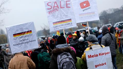 Mehrere Personen stehen bei einer Querdenken-Demonstration im Freien. Sie halten Plakate mit Aufschriften wie „Mein Körper gehört mir“ und „Grundgesetz ist keine Verschwörungstheorie“. Im Hintergrund ist eine Deutschlandfahne zu sehen.