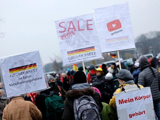 Mehrere Personen stehen bei einer Querdenken-Demonstration im Freien. Sie halten Plakate mit Aufschriften wie „Mein Körper gehört mir“ und „Grundgesetz ist keine Verschwörungstheorie“. Im Hintergrund ist eine Deutschlandfahne zu sehen.