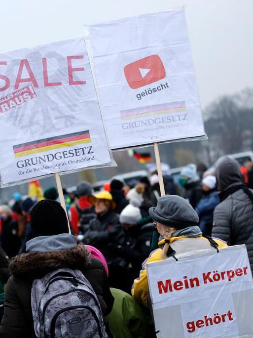 Mehrere Personen stehen bei einer Querdenken-Demonstration im Freien. Sie halten Plakate mit Aufschriften wie „Mein Körper gehört mir“ und „Grundgesetz ist keine Verschwörungstheorie“. Im Hintergrund ist eine Deutschlandfahne zu sehen.