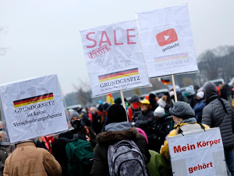Mehrere Personen stehen bei einer Querdenken-Demonstration im Freien. Sie halten Plakate mit Aufschriften wie „Mein Körper gehört mir“ und „Grundgesetz ist keine Verschwörungstheorie“. Im Hintergrund ist eine Deutschlandfahne zu sehen.