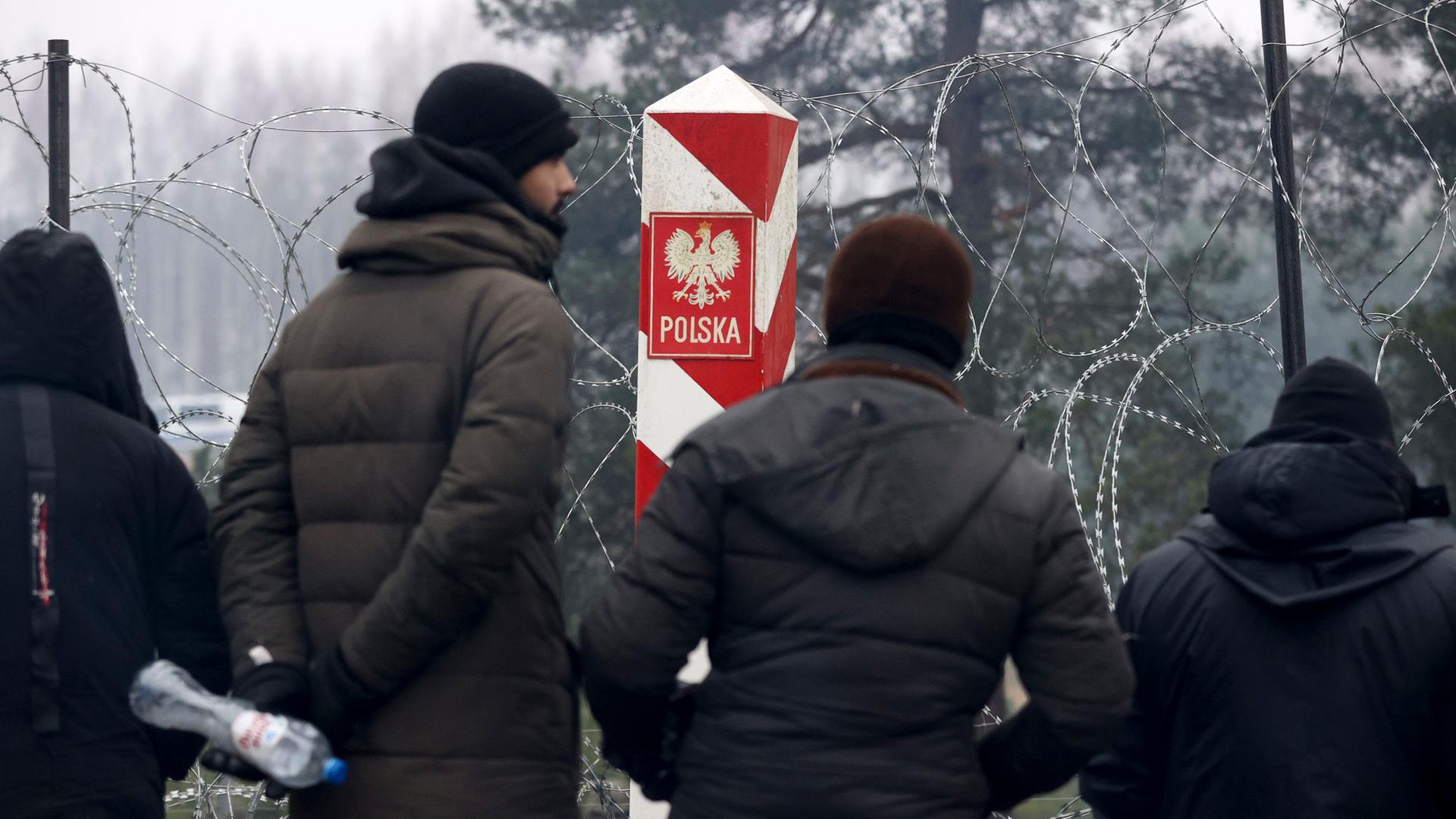 Migranten stehen in vor einem Stacheldrahtzauns und einem polnischen Grenzpfeiler an der belarussisch-polnischen Grenze in der Nähe von Grodno im  November 2021.