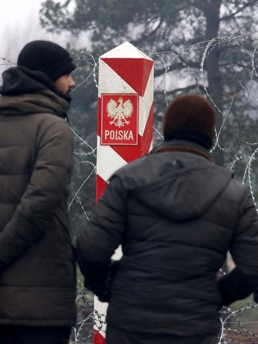 Migranten stehen in vor einem Stacheldrahtzauns und einem polnischen Grenzpfeiler an der belarussisch-polnischen Grenze in der Nähe von Grodno im  November 2021.