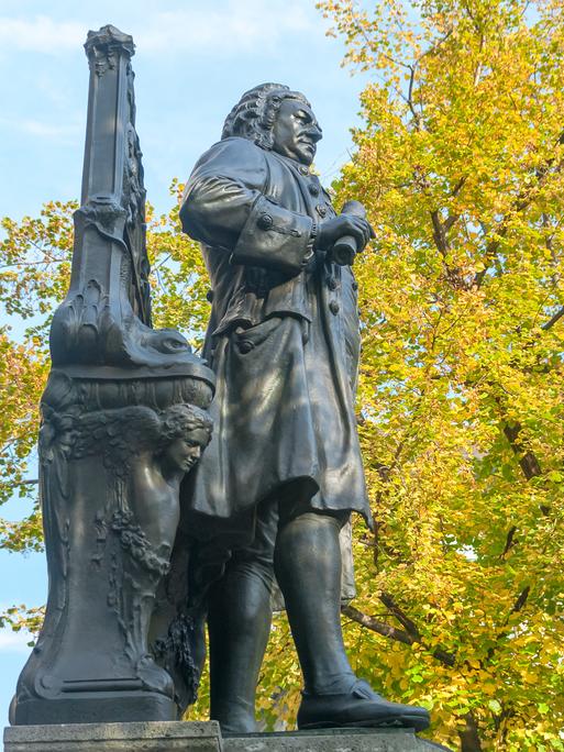 Denkmal Bachs vor der Leipziger Thomaskirche von der Seite fotografiert, sodass die Figur vor Herbslaub erscheint.