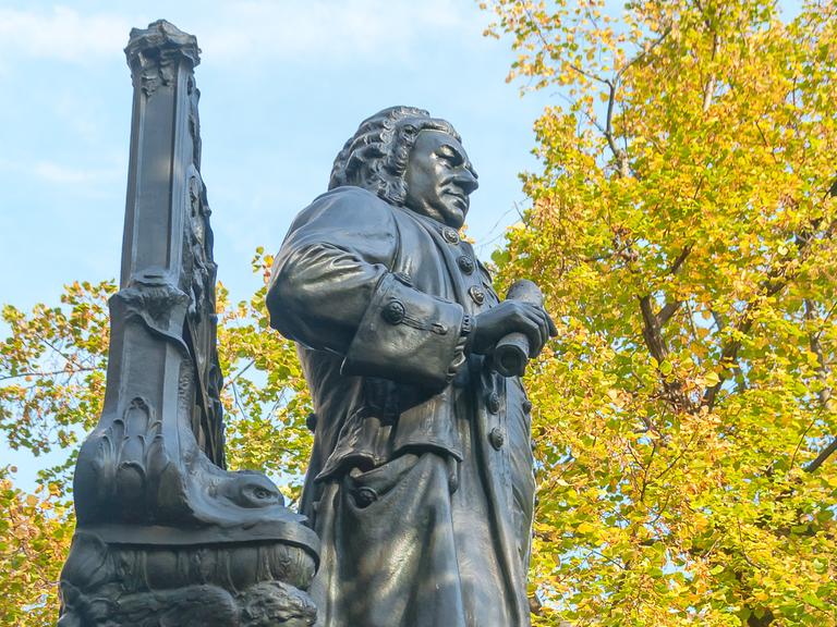 Denkmal Bachs vor der Leipziger Thomaskirche von der Seite fotografiert, sodass die Figur vor Herbslaub erscheint.