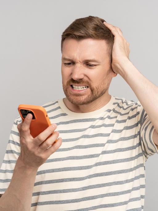 Ein junger Mann schaut mit erschrockener Miene auf sein Handy und kratzt sich am Kopf