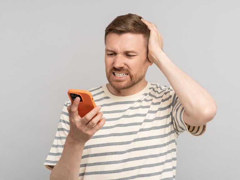 Ein junger Mann schaut mit erschrockener Miene auf sein Handy und kratzt sich am Kopf