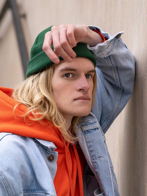 Frustriert schauender Mann mit grüner Mütze, orangen Hoodie und Jeansjacke lehnt an Wand