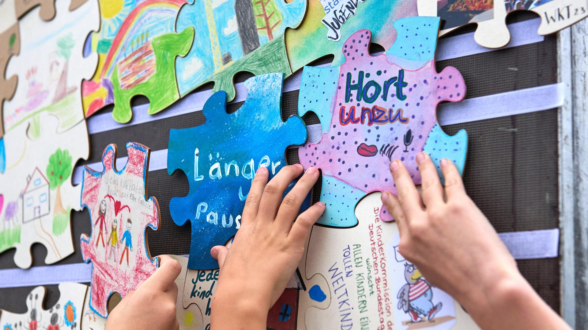 Verschiedene Puzzlestücke mit Forderungen von Kindern, wie "Mehr Pausen" oder "Hört uns zu" hängen an einer Wand. Kinderhände bringen die Puzzlestücke an. 