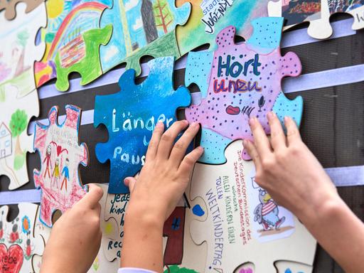 Verschiedene Puzzlestücke mit Forderungen von Kindern, wie "Mehr Pausen" oder "Hört uns zu" hängen an einer Wand. Kinderhände bringen die Puzzlestücke an. 