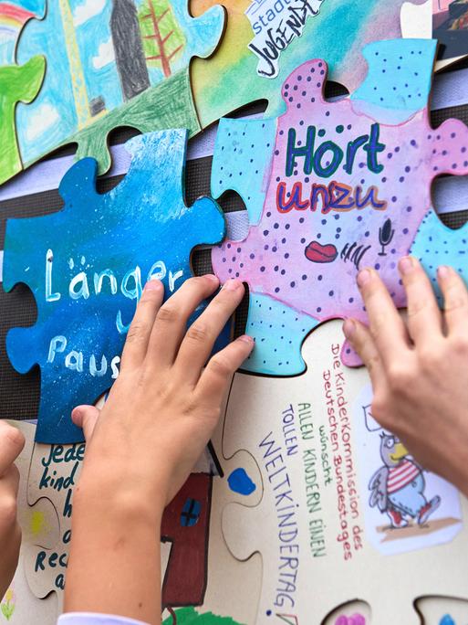 Verschiedene Puzzlestücke mit Forderungen von Kindern, wie "Mehr Pausen" oder "Hört uns zu" hängen an einer Wand. Kinderhände bringen die Puzzlestücke an. Verschiedene Puzzlestücke mit Forderungen von Kindern, wie "Mehr Pausen" oder "Hört uns zu" hängen an einer Wand. Kinderhände bringen die Puzzlestücke an.