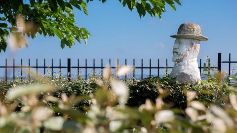 Eine steinerne Figur von Erik Satie mit Hut blickt inmitten von Grün auf das Meer. 