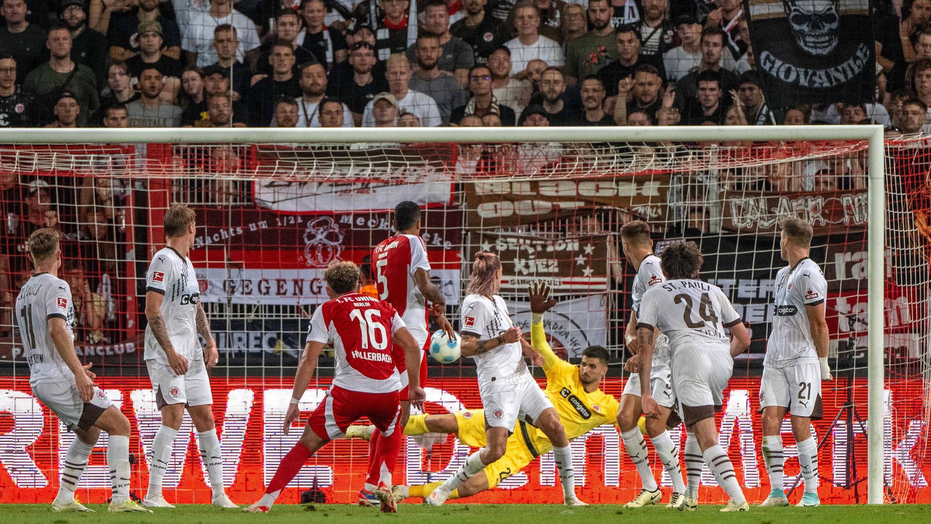 Szene aus dem Bundesliga-Spiel Union Berlin gegen St. Pauli; ein Spieler von Union Berlin schießt den Ball auf das gegnerische Tor. Spieler von St.Pauli sind zu sehen; der Torwart von St. Pauli springt in die linke Ecke.