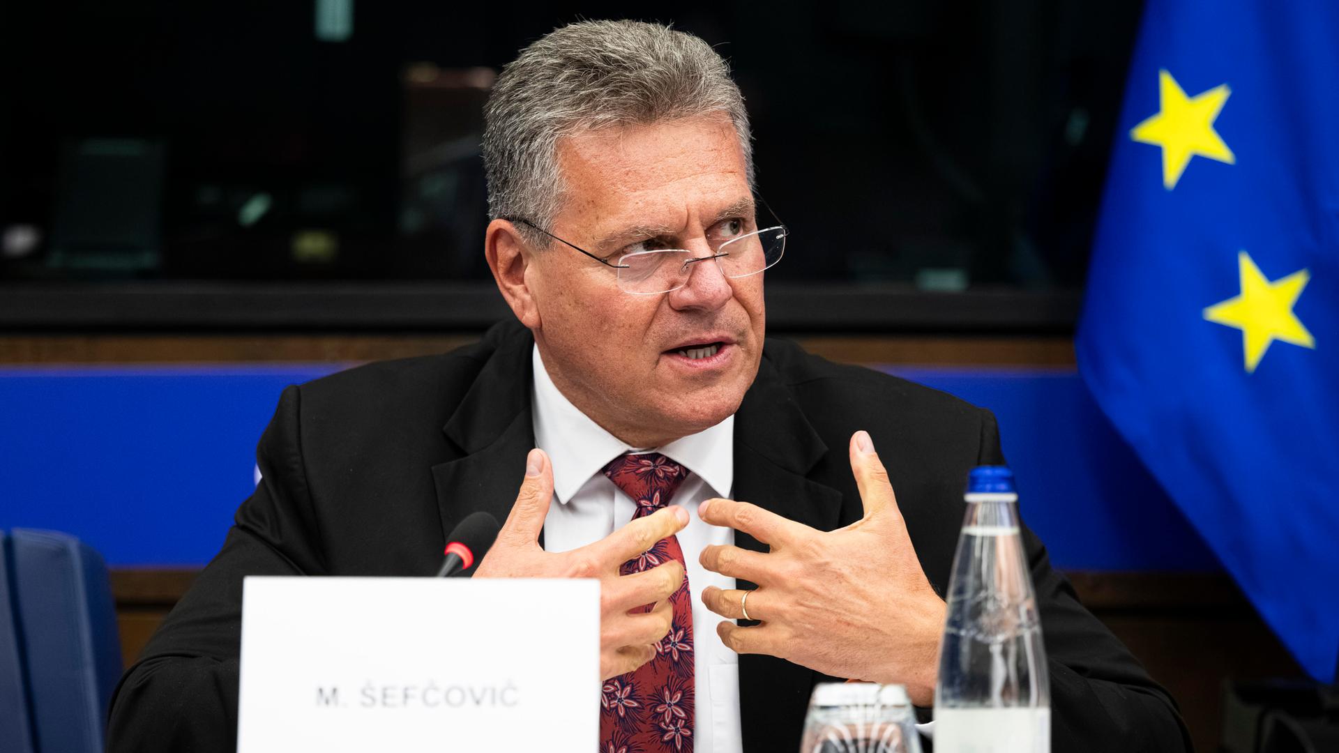 Handelskommissar Maroš Šefčovič bei einer EU-Veranstaltung. Er trägt ein blaues Jackett. Er gestikuliert mit den Händen und trägt eine Brille, die etwas von der Nase gerutscht ist. Vor ihm steht ein Schild mit seinem Namen.
