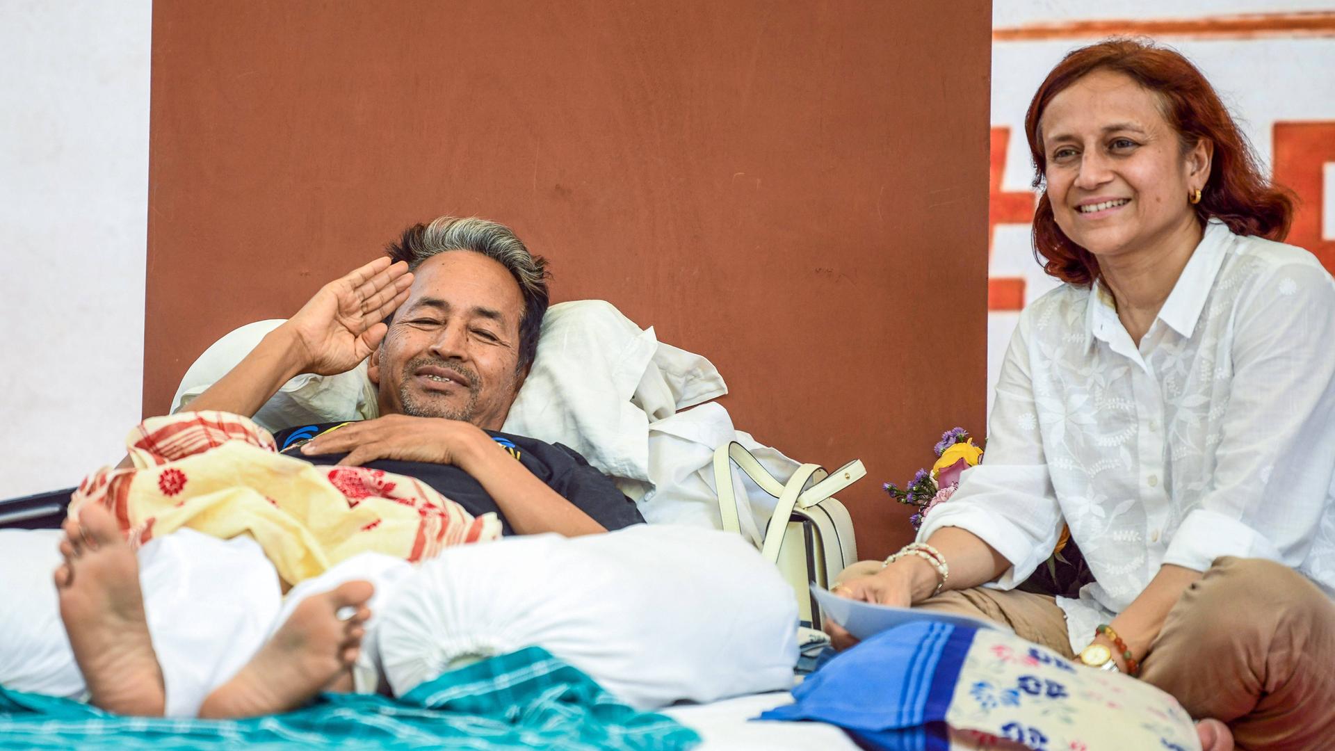 Der indische Aktivist Sonam Wangchuk liegt im Hungerstreik in einem Bett und winkt in Richtung Kamera. Neben im sitzt eine lächelnde Frau.
Der indische Aktivist Sonam Wangchuk liegt im Hungerstreik in einem Bett und winkt in Richtung Kamera. Neben im sitzt eine lächelnde Frau.