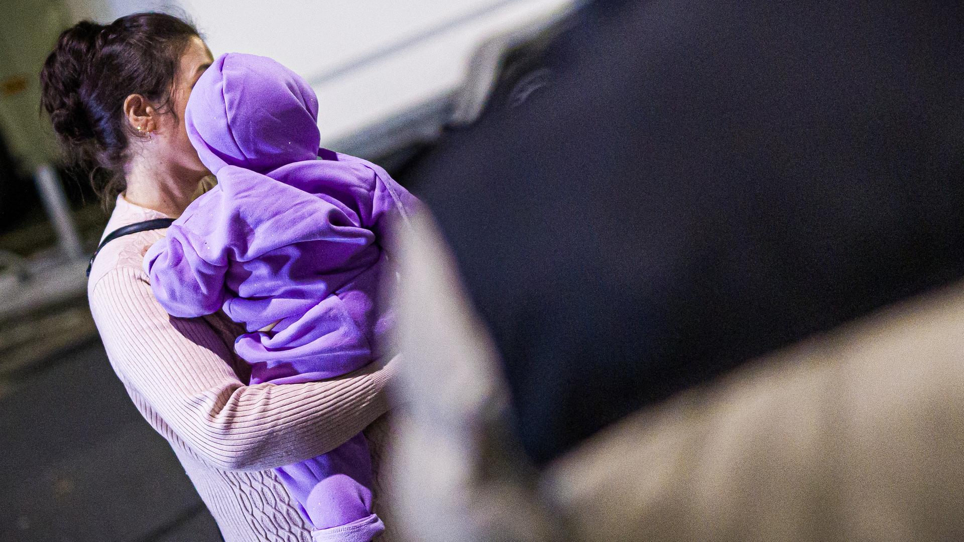 Eine Frau trägt ein Kleinkind auf dem Arm. Ihr Gesicht wird durch das Kind verdeckt, das eine Kapuze trägt und von hinten fotografiert wurde. 