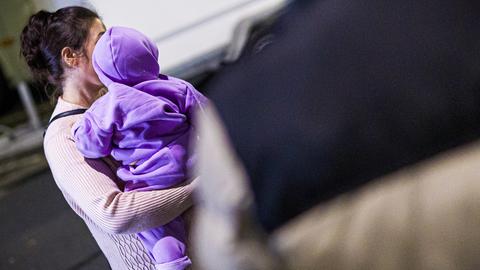 Eine Frau trägt ein Kleinkind auf dem Arm. Ihr Gesicht wird durch das Kind verdeckt, das eine Kapuze trägt und von hinten fotografiert wurde. 