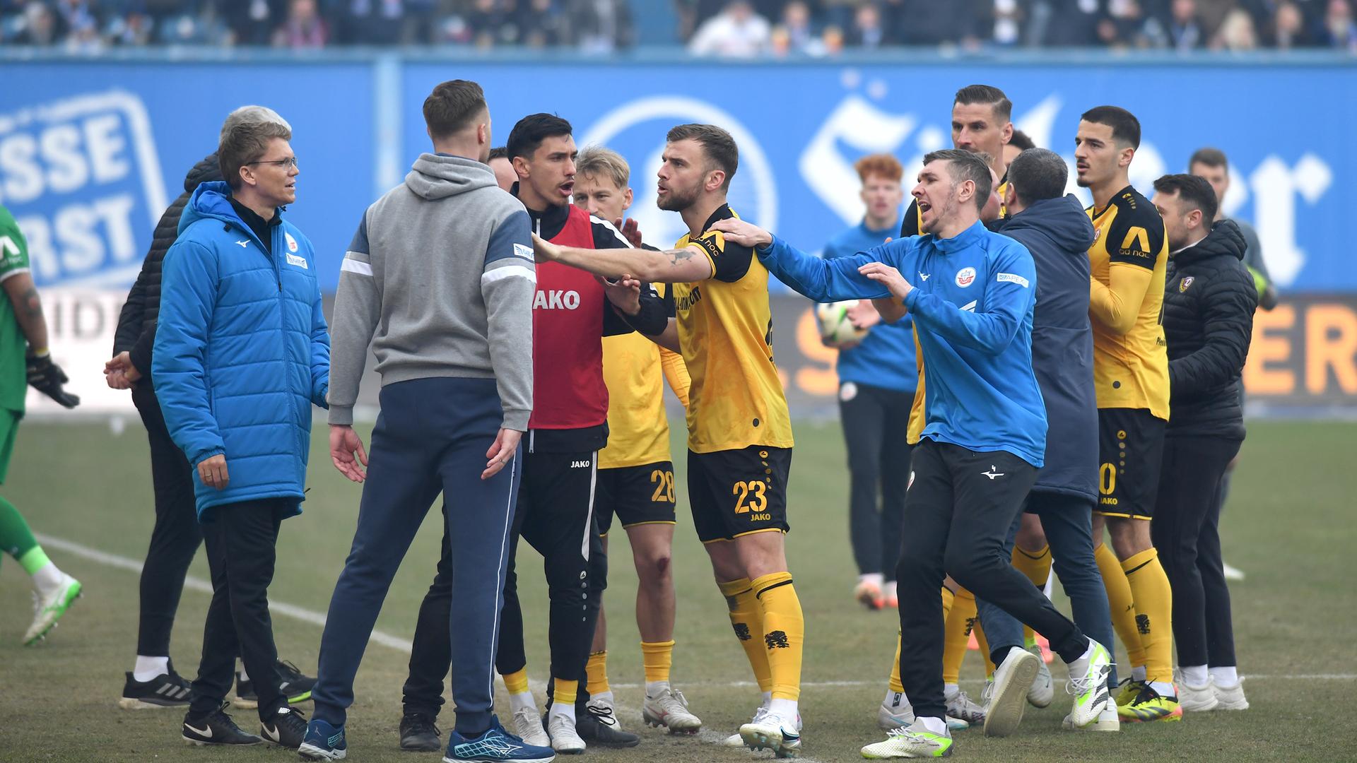 Auf einem Fußballplatz stößt ein offenbar wütender Spieler einem Verantwortlichen vor die Brust. Andere Spieler versuchen, ihn zurückzuhalten. 