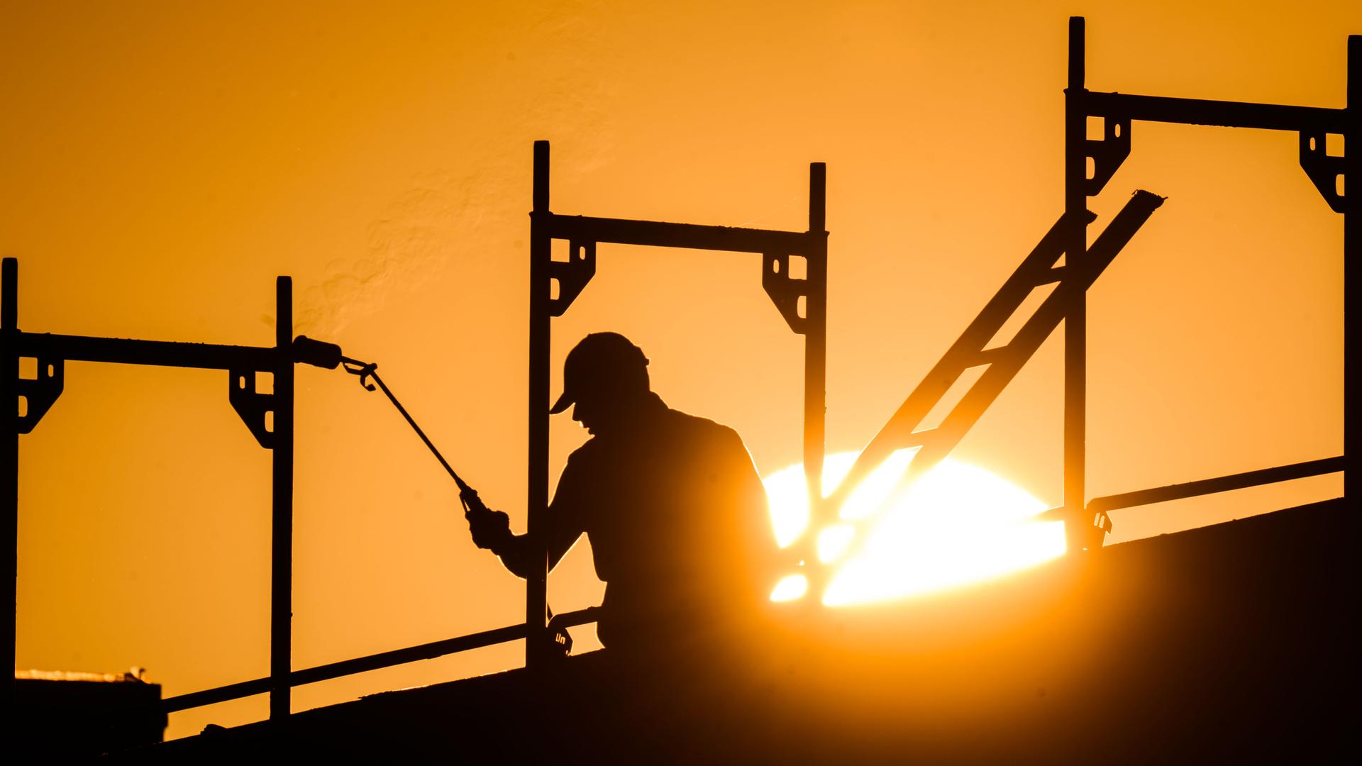 Ein Bauarbeiter steht mit einem Gasbrenner beim Bau eines Mehrfamilienhauses im Neubaugebiet Kronsrode, als am Horizont die Sonne aufgeht.