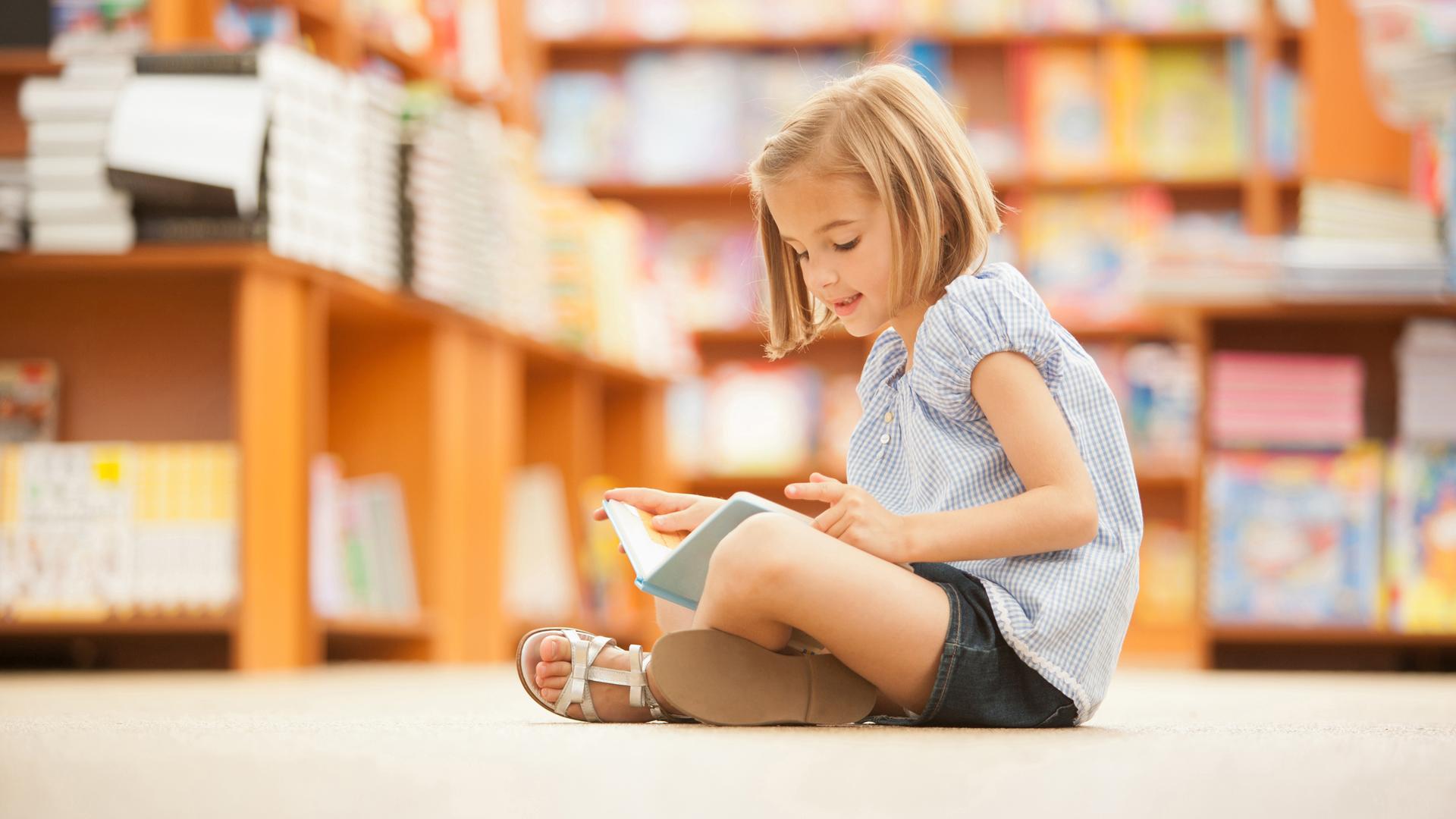 Ein Mädchen sitzt mit einem Buch in der Hand in einer Bibliothek auf dem Boden und liest.
