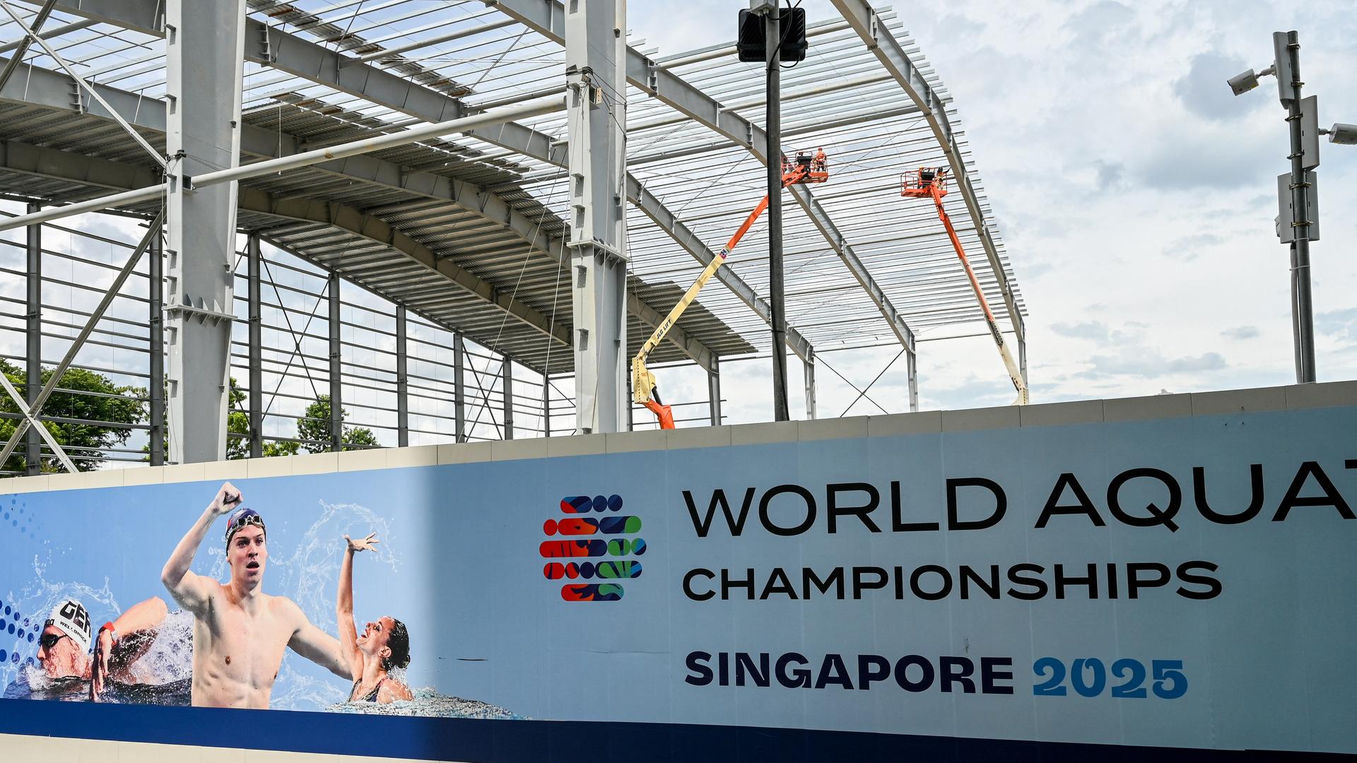 Vor einem noch im Bau befindlichen Stadion hängt ein Plakat mit der Aufschrift "World Aquatics Championships Singapore 2025". Darauf sind Vertreterinnen und Vertreter verschiedener Schwimm-Disziplinen abgebildet. Vor einem noch im Bau befindlichen Stadion hängt ein Plakat mit der Aufschrift "World Aquatics Championships Singapore 2025". Darauf sind Vertreterinnen und Vertreter verschiedener Schwimm-Disziplinen abgebildet.
