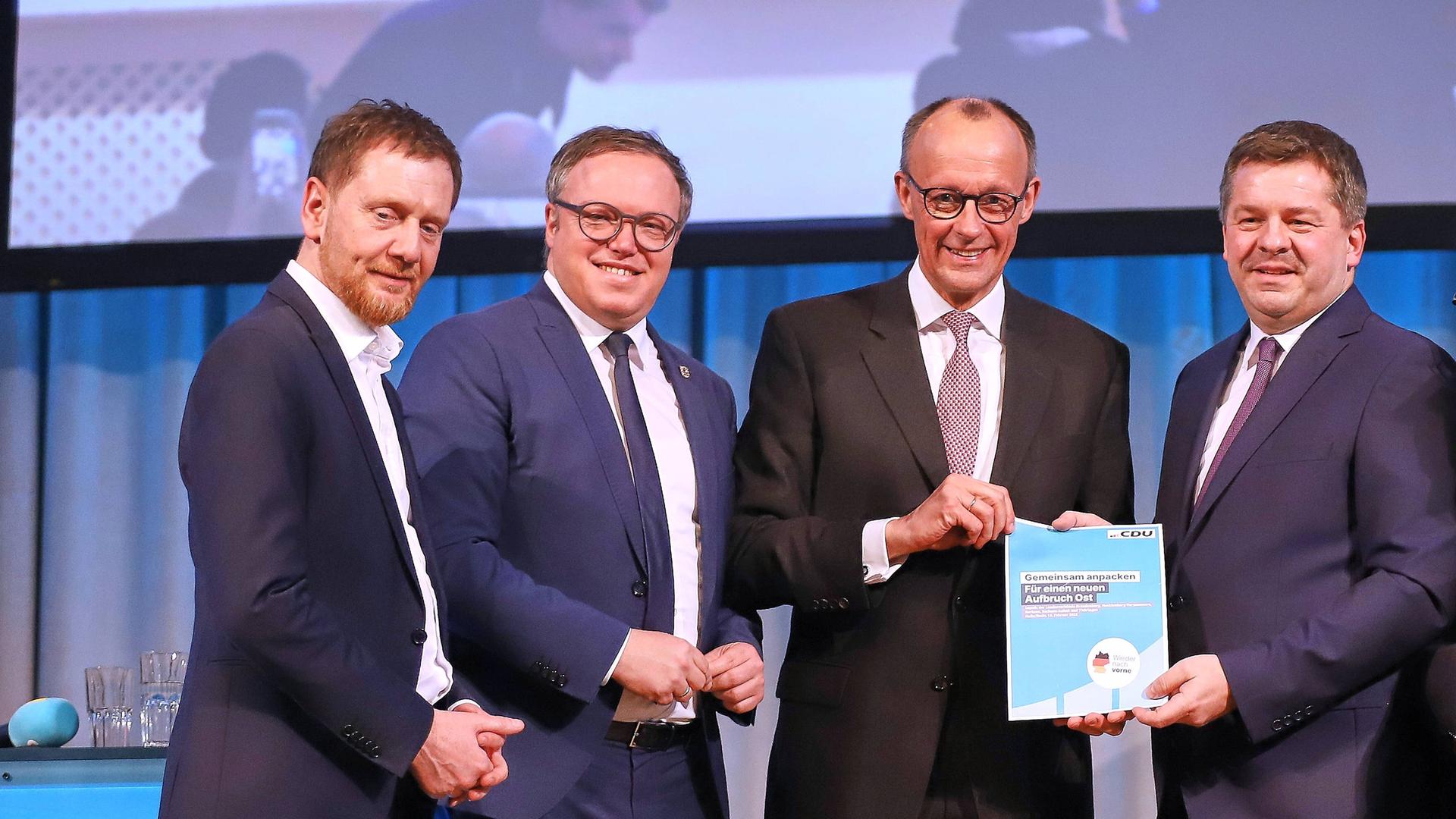 Michael Kretschmer, Mario Voigt, Friedrich Merz und Sven Schulze stehen nebeneinander auf einer Bühne. Sie schauen in Richtung der Kamera.