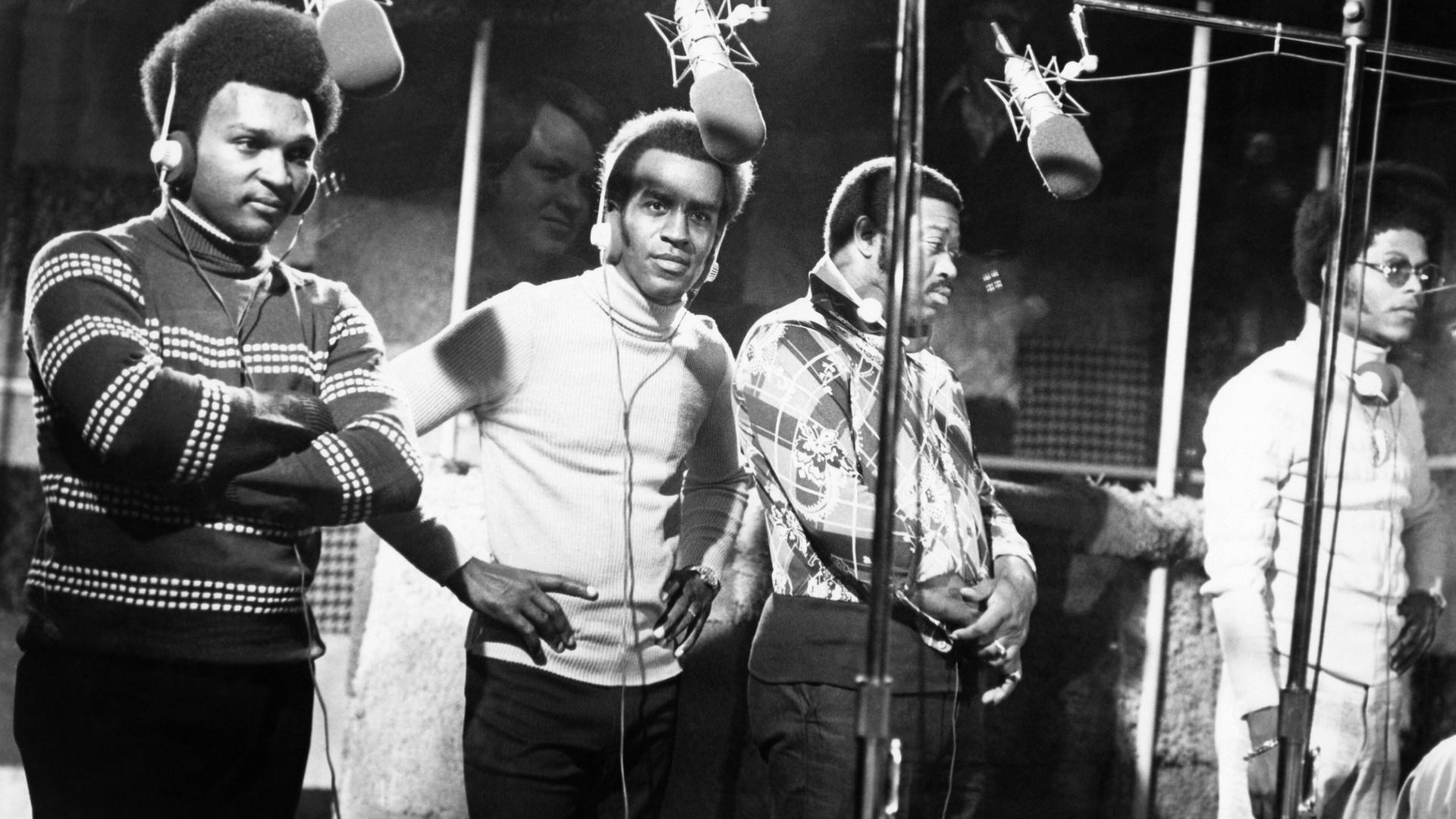 Schwarz-weiß-Foto der Band The Impressions. Vier Männer mit Kopfhörern stehen in einem Studio vor Mikrofoene.