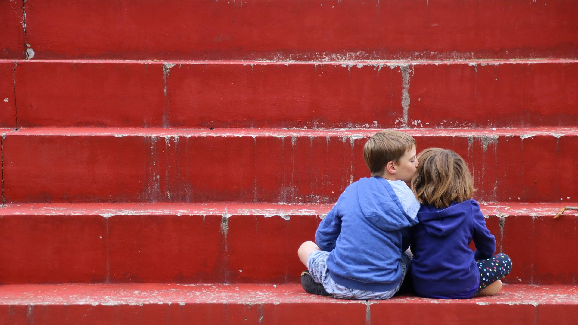 Zwei Kinder in Kapuzenpullis sitzen auf einer roten Treppe mit dem Rücken zu den Betrachtenden. Das linke Kind küsst das rechte Kind auf die Wange.