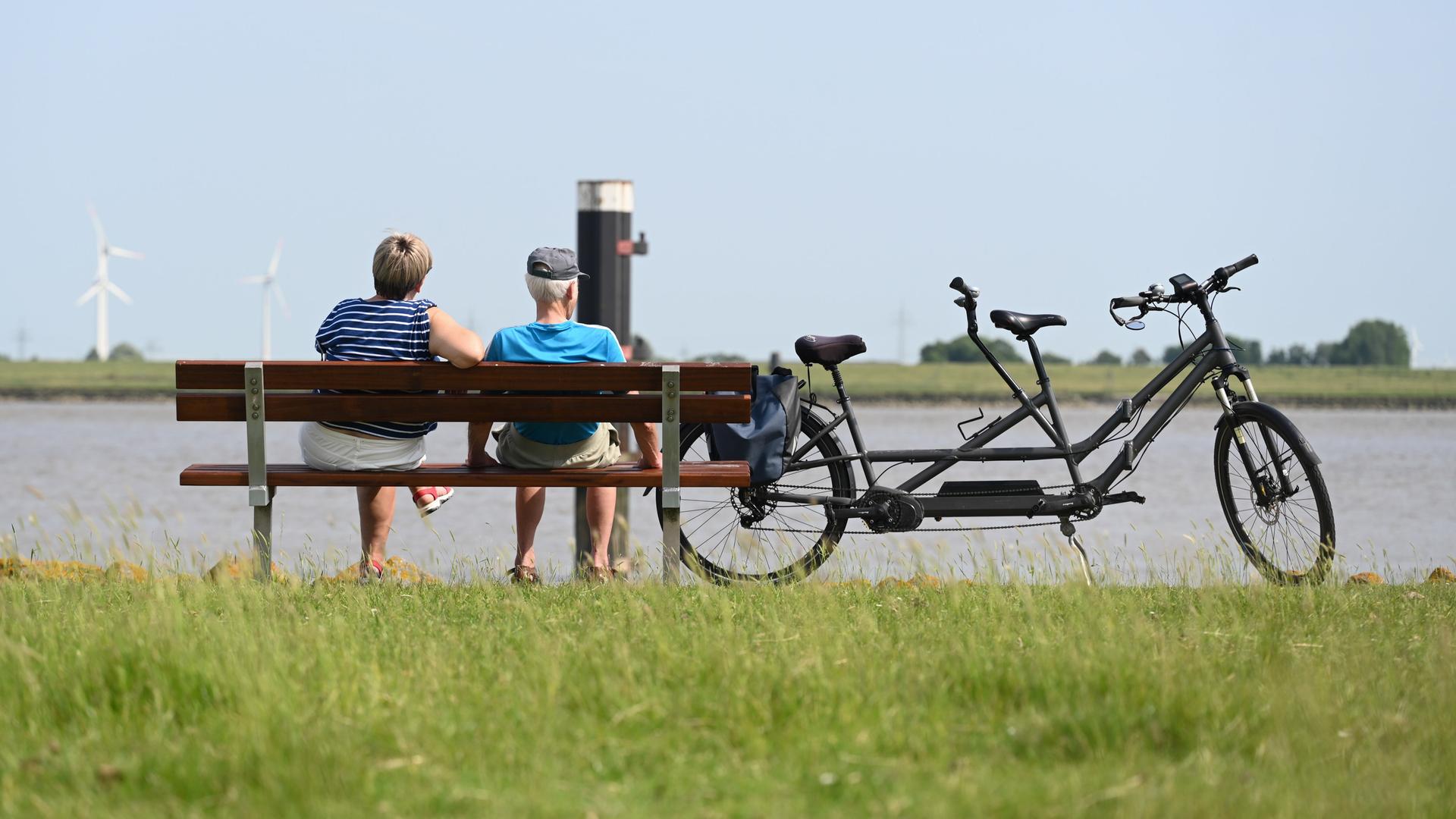 Zwei Personen sitzen bei strahlendem Sommerwetter neben ihrem Tandem auf einer Bank am Deich
