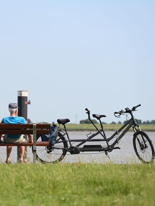 Zwei Personen sitzen bei strahlendem Sommerwetter neben ihrem Tandem auf einer Bank am Deich