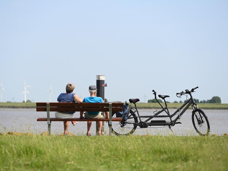 Zwei Personen sitzen bei strahlendem Sommerwetter neben ihrem Tandem auf einer Bank am Deich
