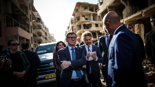 Ein Mann steht vor zerstörten Gebäuden und hört einen Gesprächspartner zu.