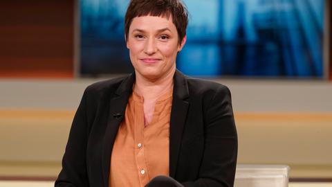 Nicole Deitelhoff sitzt im Fernsehstudio von Anne Will, trägt ein schwarzes Sacko und eine organgefarbene Bluse und schaut freundlich in die Kamera