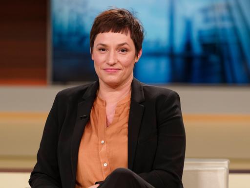 Nicole Deitelhoff sitzt im Fernsehstudio von Anne Will, trägt ein schwarzes Sacko und eine organgefarbene Bluse und schaut freundlich in die Kamera