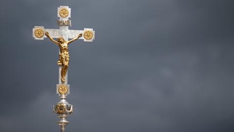Ein goldenes Kruzifix ist vor dunklen Wolken zu sehen.