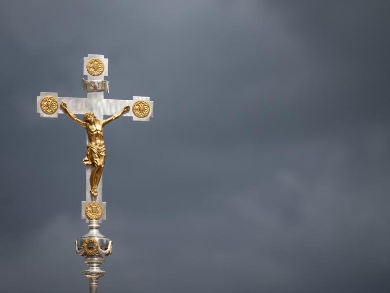 Ein goldenes Kruzifix ist vor dunklen Wolken zu sehen.