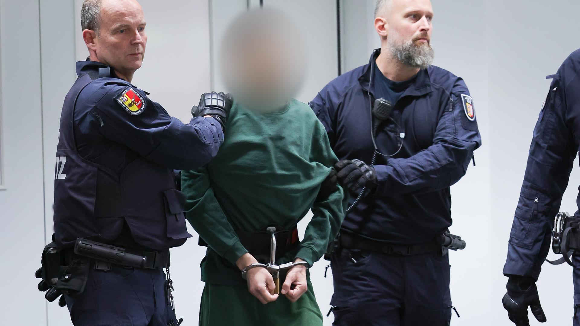 Zwei Polizisten bringen den Angeklagten in Handschellen, der in grüner Haftkeldung erscheint. Sein Gesicht ist unscharf gemacht. 