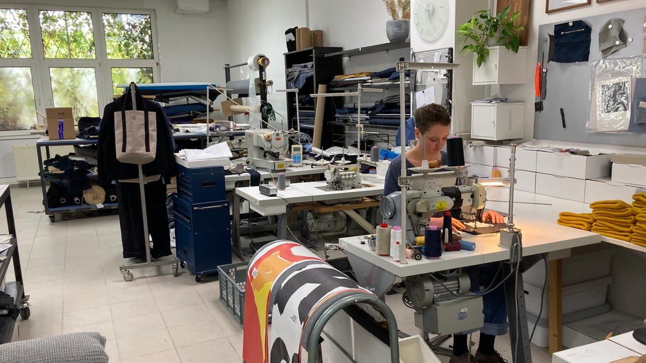 Die Schneiderin und Schnittmacherin Catharina Saffier sitzt in ihrem Berliner Atelier an einer Nähmaschine, im Hintergrund stehen weitere Maschinen und Ständer mit Stoffen und Kleidunggsstücken.