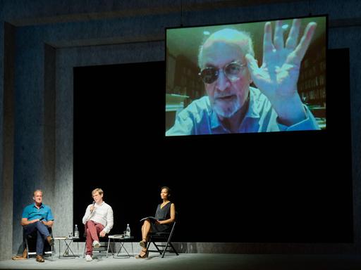 Schriftsteller Salman Rushdie zeigt, live aus New York zugeschaltet, seine bei einem Anschlag verletzte und inzwischen genesene Hand, während der Übersetzer Bernhard Robben, Schriftsteller Daniel Kehlmann und Schauspielerin Cynthia Micas der Bühne sitzen. 