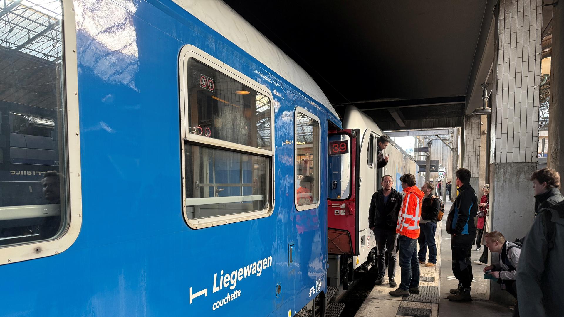 Menschen stehen auf einem Bahnsteig vor einem Zug. Auf dem Waggon steht das Wort "Liegewagen". Menschen stehen auf einem Bahnsteig vor einem Zug. Auf dem Waggon steht das Wort "Liegewagen".
