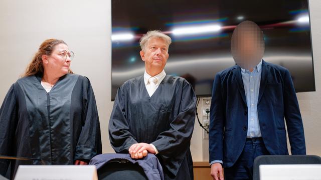 Der Angeklagte steht im Verhandlungs-Saal von dem Land-Gericht neben seinen Rechtsanwälten. Sein Gesicht ist nicht zu erkennen. 