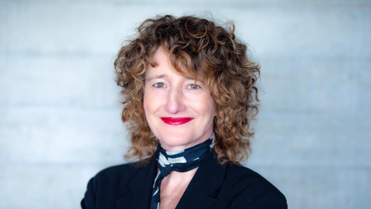 Eine Frau mit dunkelblonden Locken schaut freundlich lächelnd in die Kamera. Sie hat ein schwarzes Oberteil und einen schwarz-weißen Schal an. Ihre Lippen sind rot geschminkt. 