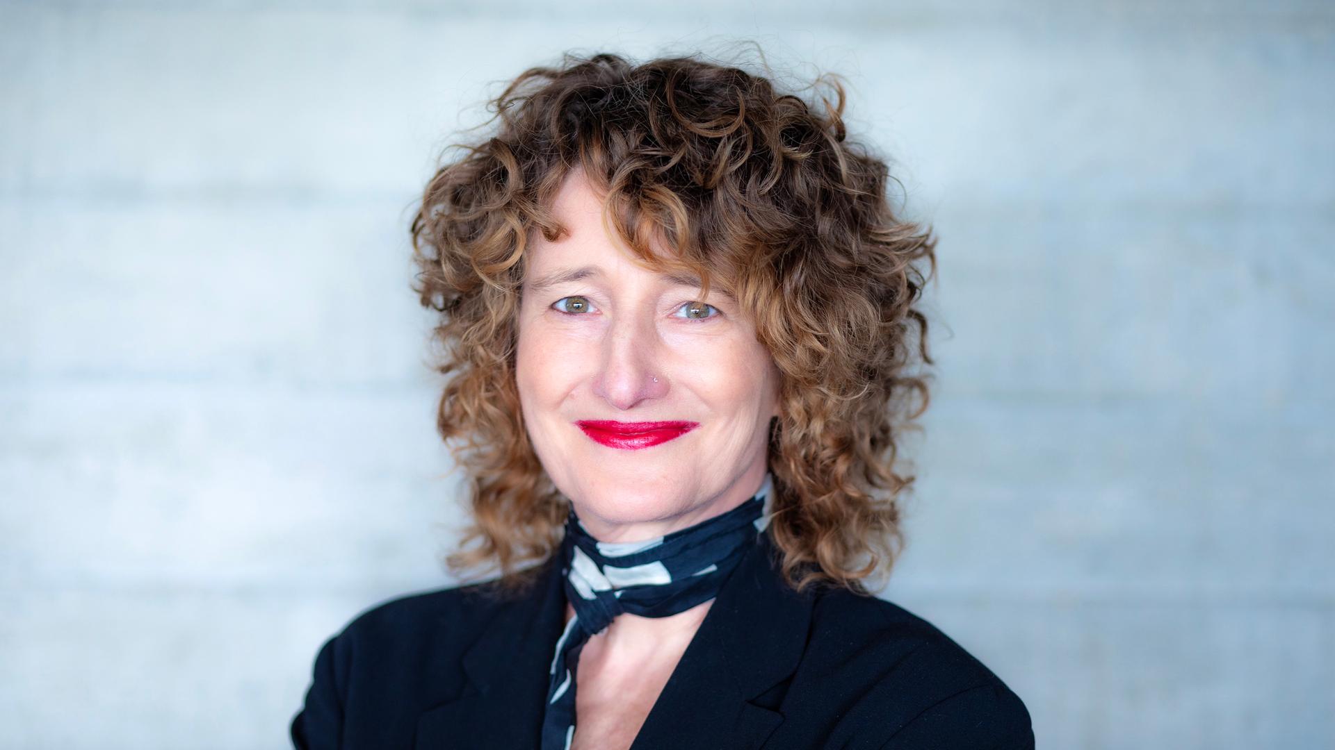 Eine Frau mit dunkelblonden Locken schaut freundlich lächelnd in die Kamera. Sie hat ein schwarzes Oberteil und einen schwarz-weißen Schal an. Ihre Lippen sind rot geschminkt. 