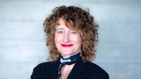 Eine Frau mit dunkelblonden Locken schaut freundlich lächelnd in die Kamera. Sie hat ein schwarzes Oberteil und einen schwarz-weißen Schal an. Ihre Lippen sind rot geschminkt. 