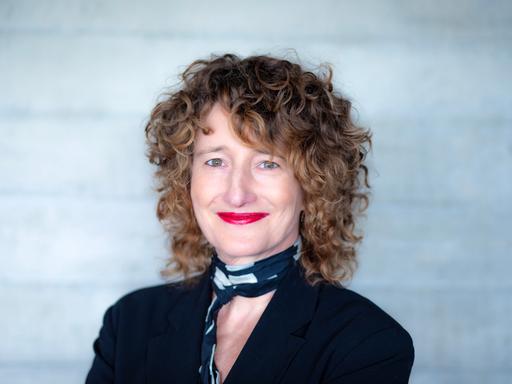 Eine Frau mit dunkelblonden Locken schaut freundlich lächelnd in die Kamera. Sie hat ein schwarzes Oberteil und einen schwarz-weißen Schal an. Ihre Lippen sind rot geschminkt. 