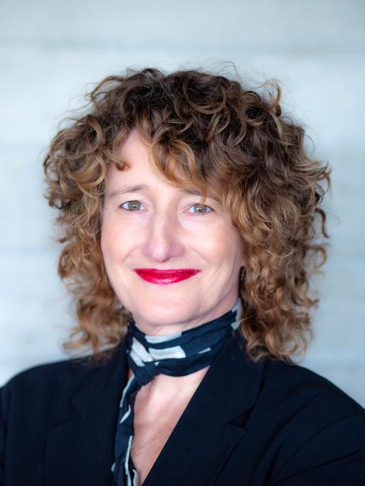 Eine Frau mit dunkelblonden Locken schaut freundlich lächelnd in die Kamera. Sie hat ein schwarzes Oberteil und einen schwarz-weißen Schal an. Ihre Lippen sind rot geschminkt. 