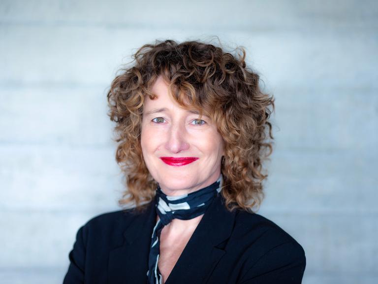 Eine Frau mit dunkelblonden Locken schaut freundlich lächelnd in die Kamera. Sie hat ein schwarzes Oberteil und einen schwarz-weißen Schal an. Ihre Lippen sind rot geschminkt. 