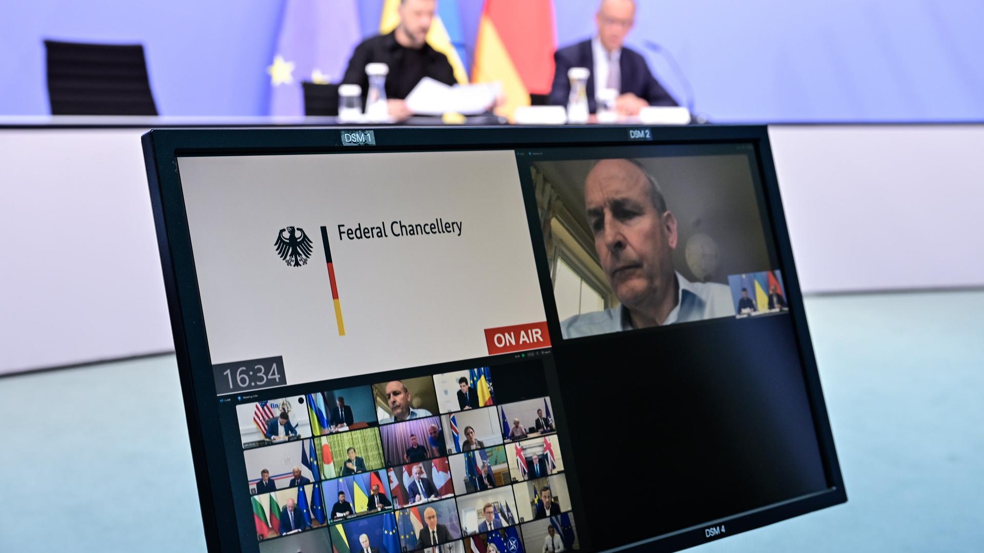 Im Vordergrund ein Bildschirm mit einer Videoschalte, deren Teilnehmer nicht erkennbar sind. Im Hintegrund klein und unscharf Merz sowie Selenskyj während der Pressekonferenz