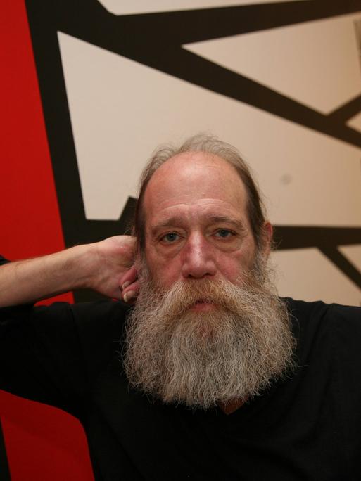 Ein Mann mit sehr langem Bart stützt seinen Kopf auf seine Faust. Hinter im sind schemenhaft große, rote Buchstaben an der Wand. Es ist Lawrence Weiner.